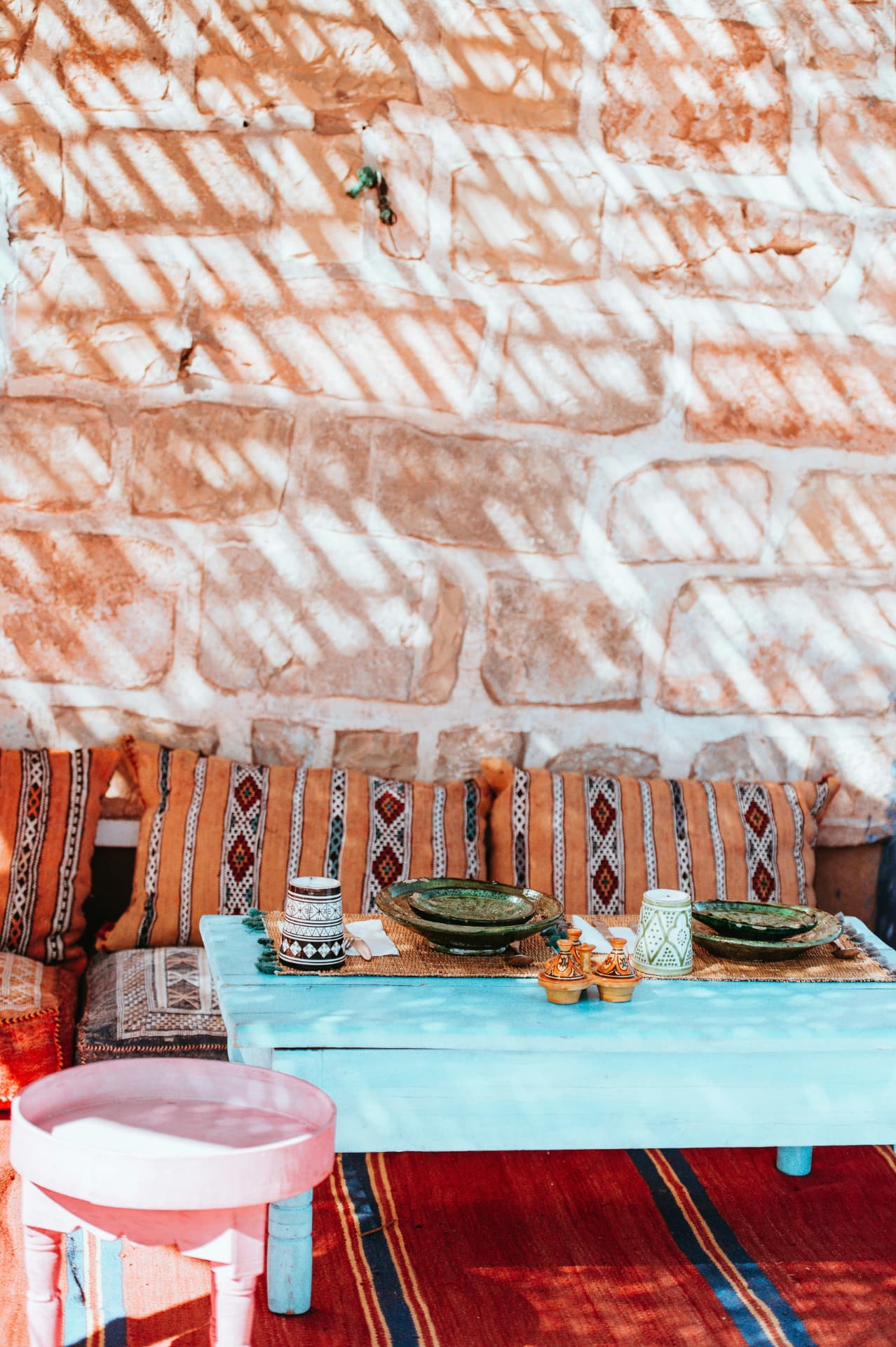 Gedekte tafel met servies, kussens en een gestreept kleed tegen een stenen muur